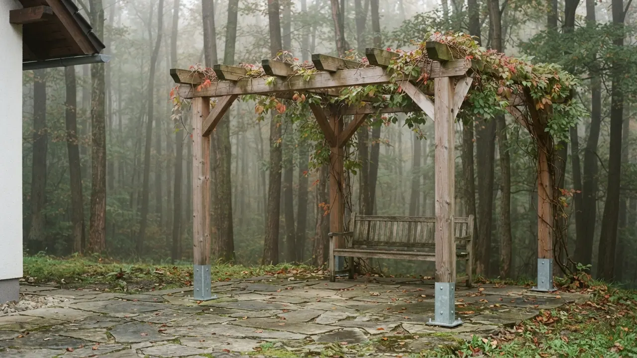 Drevená pergola nad kamennou terasou pri dome na okraji lesa, stĺpy v oceľových pätkách a trámy obrastené divým viničom v rannej hmle.